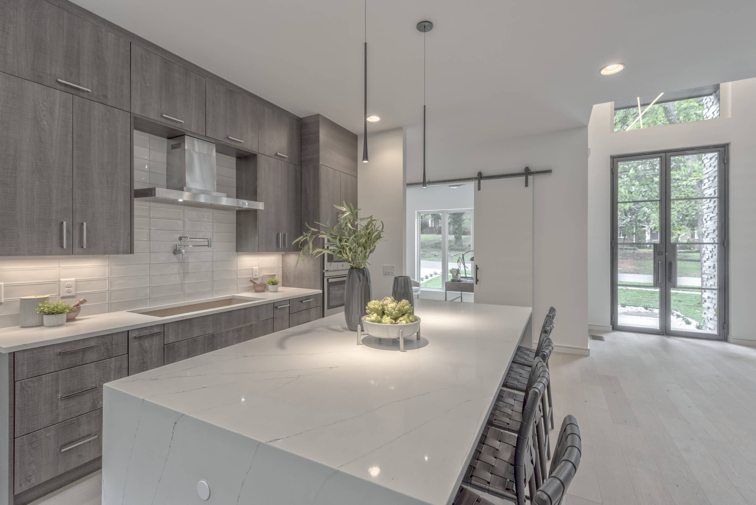Modern gray and white kitchen with wood box, dove tail drawers, and a farmhouse style sliding double door with subway tile backsplash and huge windows into forest. White marble countertops with a waterfall island