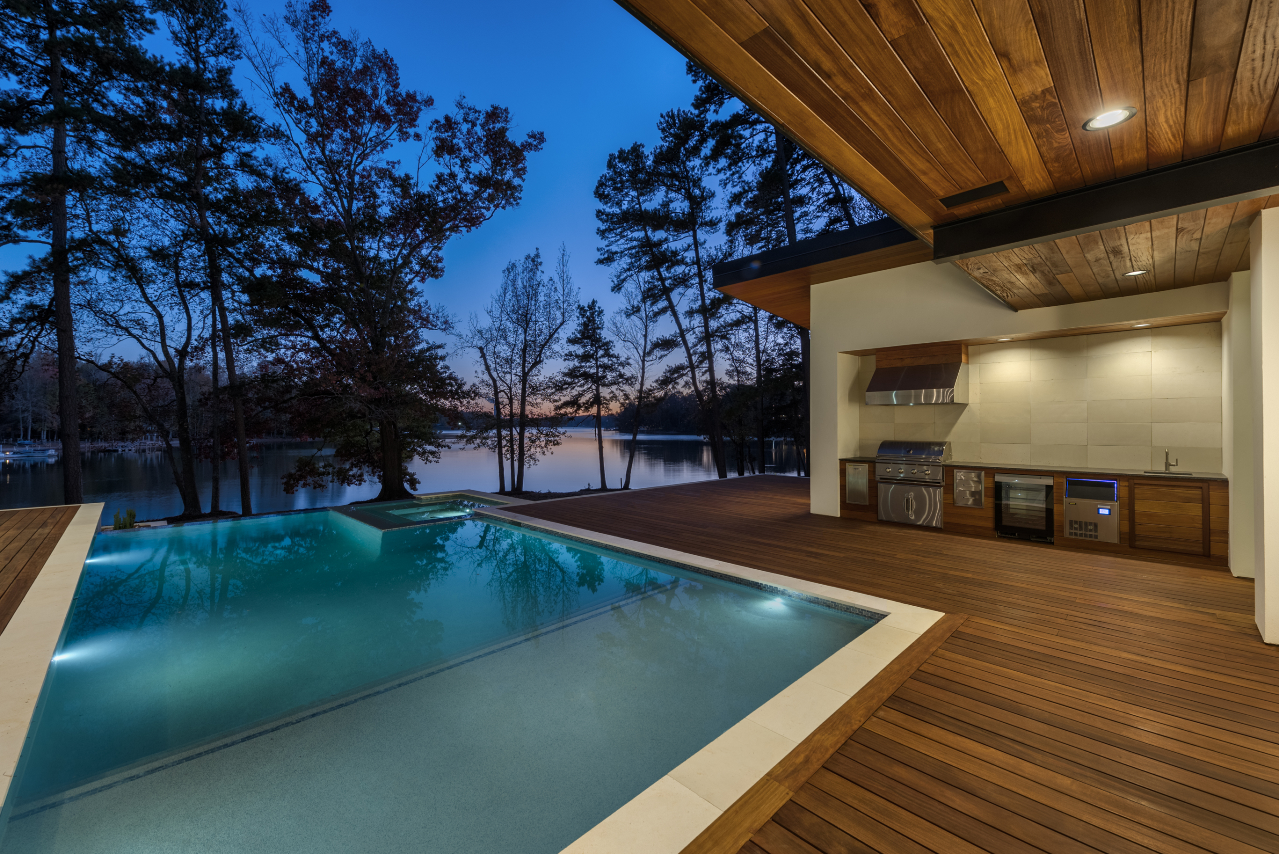 During night Cumaru wood deck and custom heated pool with infinity edge, oversized spa, tanning ledge and LED lighting package, pebble plaster finish and limestone coping and Outdoor kitchen area with built-in grill, hood, ice maker, sink and custom cabinets