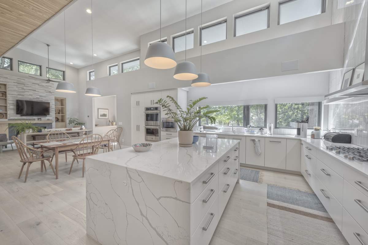 Modern white kitchen with tall ceilings and marble white countertops with waterfall island. Large windows in front of the sink. Open floor plan leads into dining room and living room