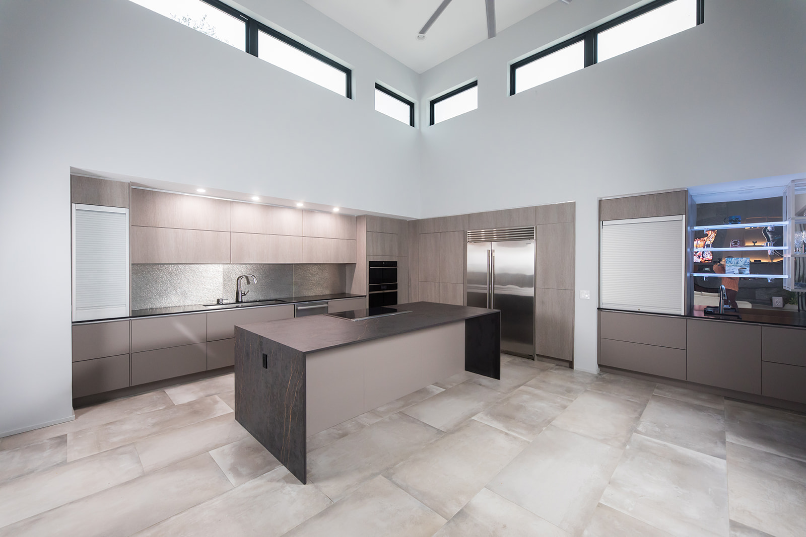 Modern gray slate minimalist kitchen with waterfall countertop, stainless steel hardware, electric flat stove, and metallic backsplash, and natural light