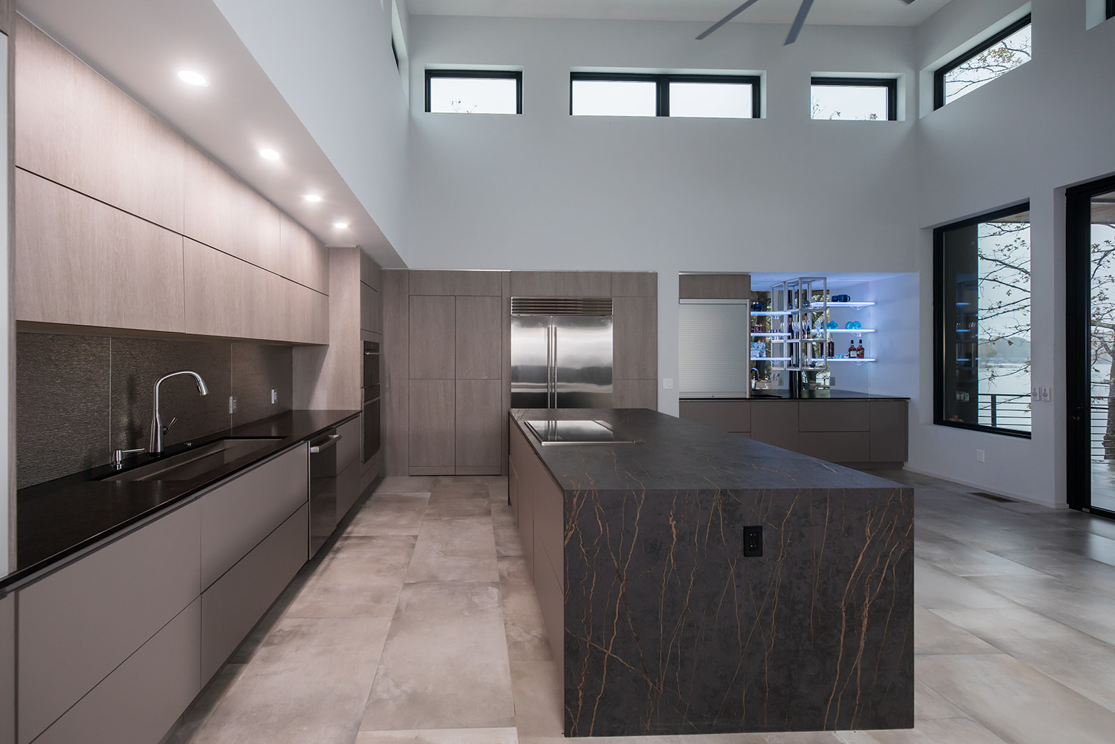 Modern gray slate minimalist kitchen with waterfall countertop, stainless steel hardware, electric flat stove, and metallic backsplash, and natural light